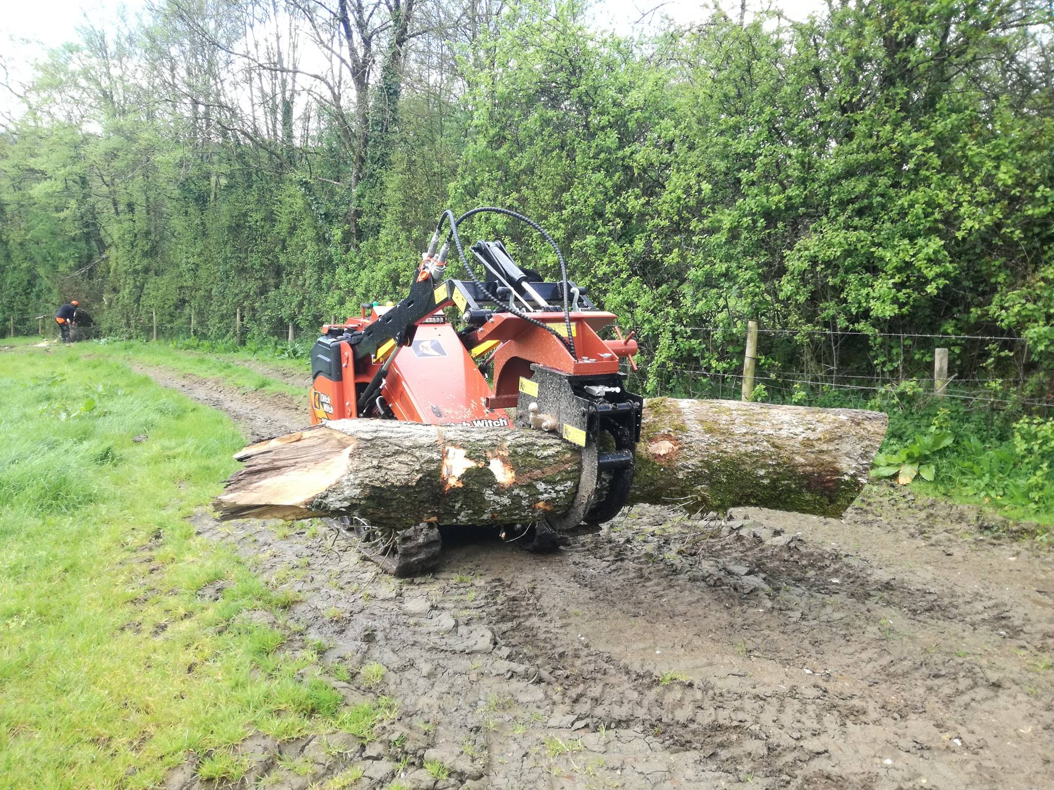 Ditch Witch tracked loader carrying a long log across a muddy field